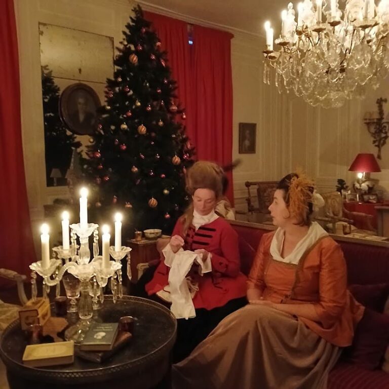 Deux figurantes costumées assises sur un canapé rouge, près du sapin, à la lueur des bougies