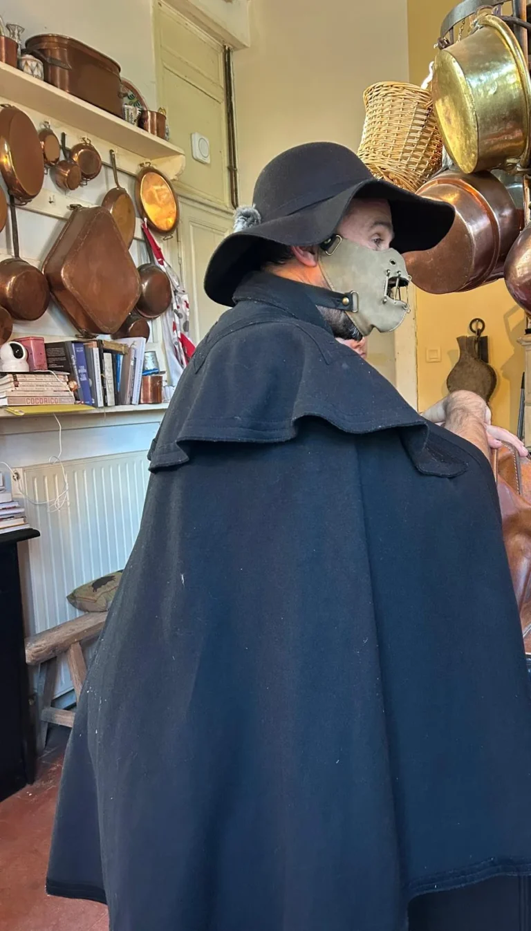 Figurant en cape noire et masque, dans la cuisine aux casseroles de cuivre